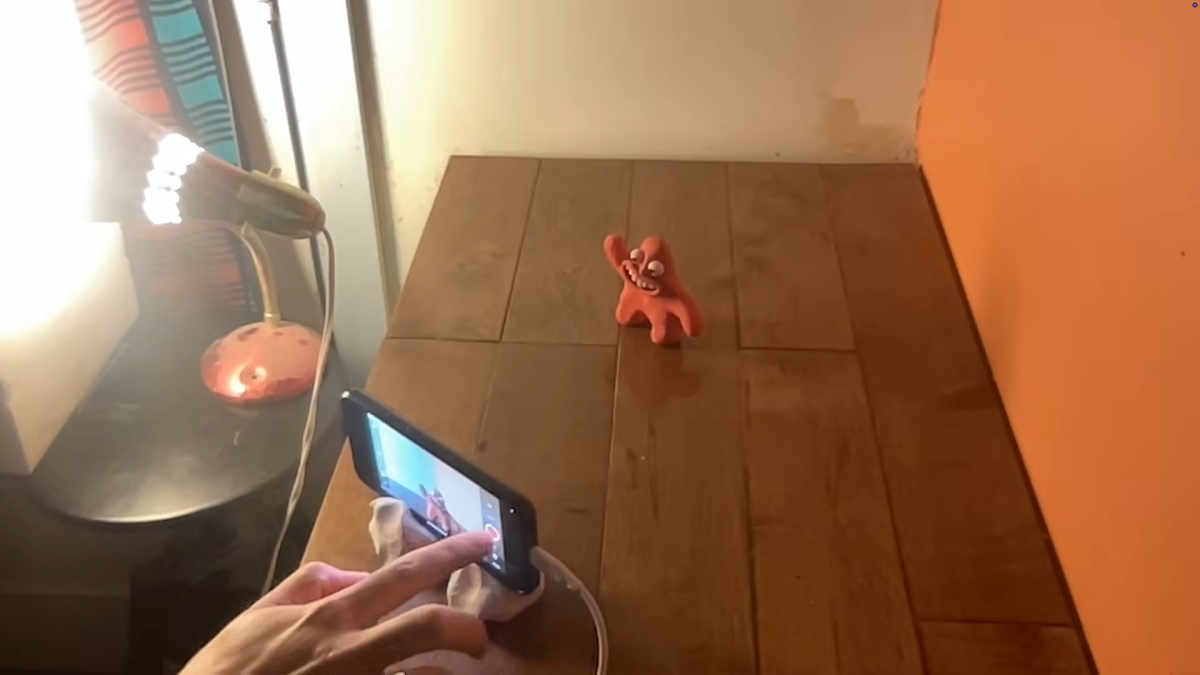 A red, cartoonish figure stands on a wooden table, appearing to wave. A smartphone held by a hand is set up on a stand, aiming to capture the figure, with a lamp and a part of a wall visible in the background.