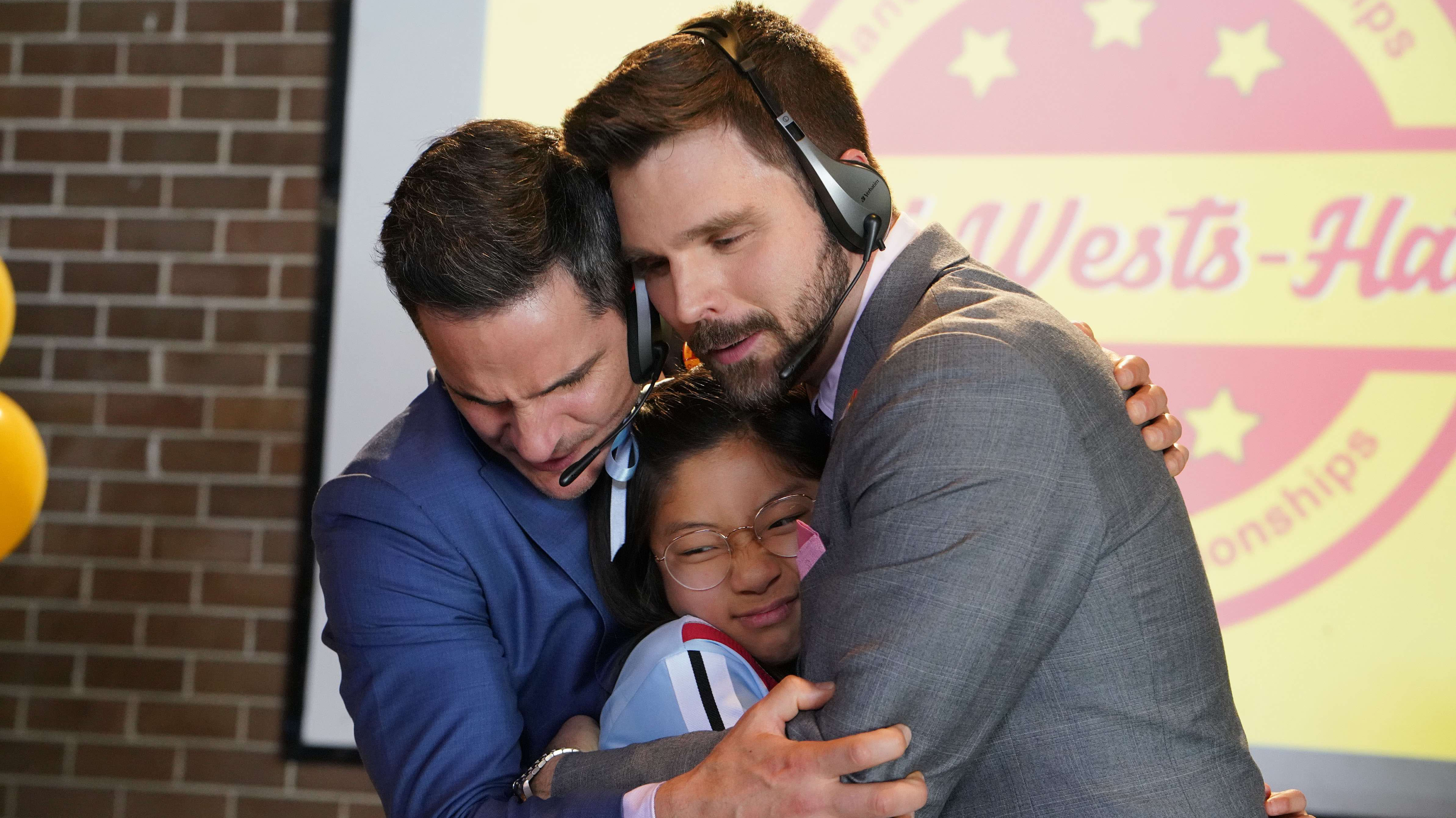 Two men wearing headsets embrace a young girl with glasses in a warm, supportive group hug. The background features a colorful logo for a championship event, suggesting a moment of celebration or achievement.