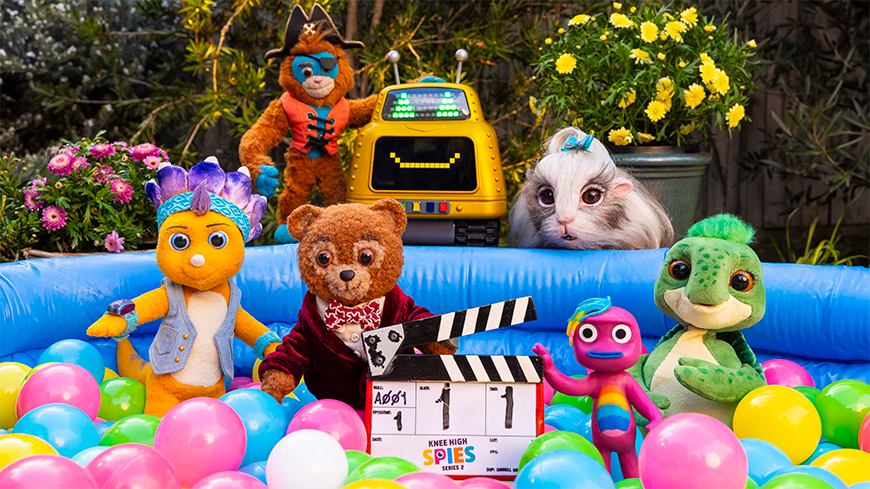 A group of colorful plush characters gathers around a blue inflatable pool filled with pastel-colored balls. Among them are a bear in a red jacket, a yellow creature with a blue mohawk, and a green crocodile, all posing cheerfully with a film clapperboard. In the background, a robot and a bunny-like creature can be seen, along with bright yellow flowers.