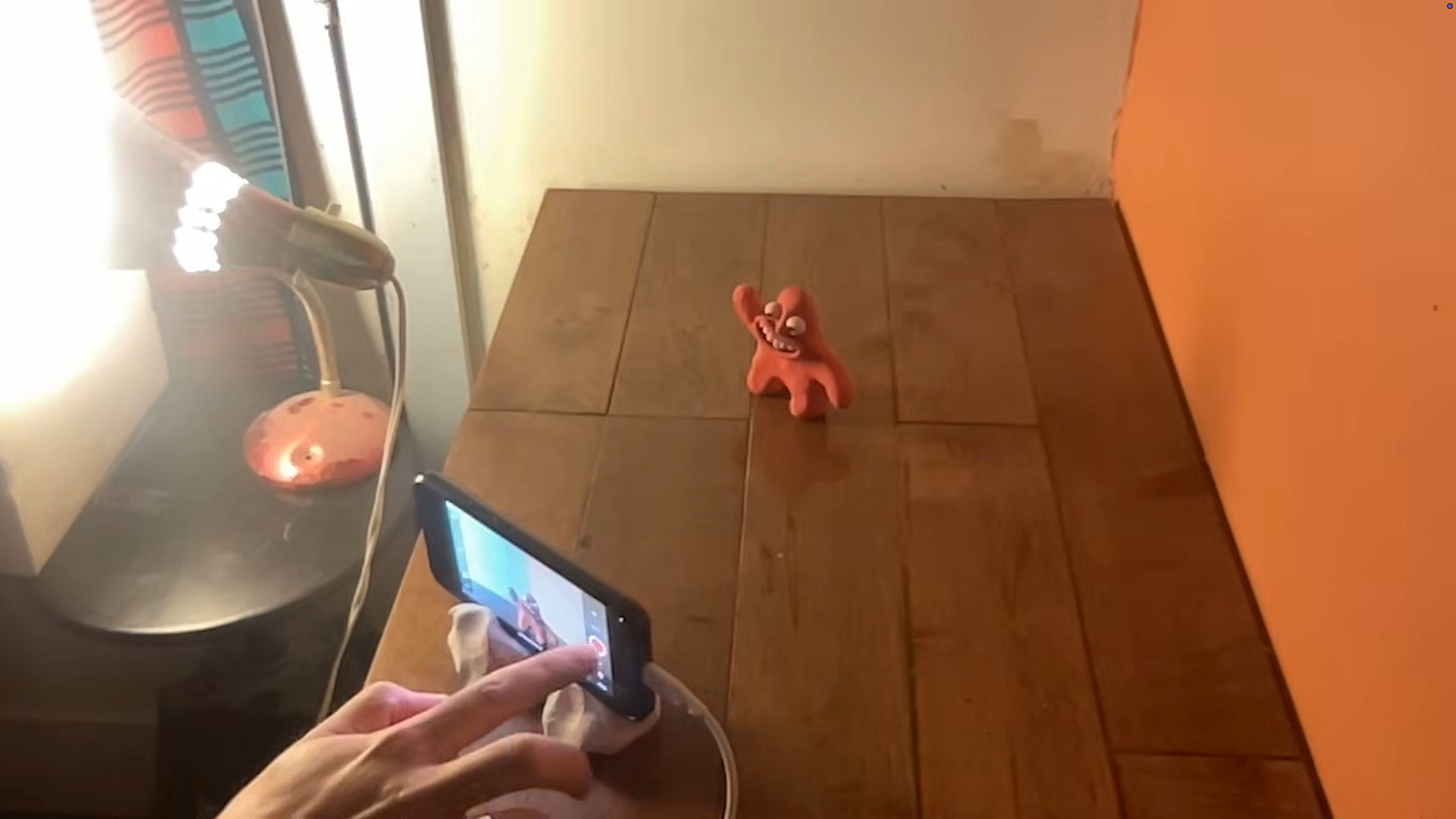 A red, cartoonish figure stands on a wooden table, appearing to wave. A smartphone held by a hand is set up on a stand, aiming to capture the figure, with a lamp and a part of a wall visible in the background.